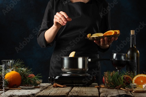 The chef pours chopped ingredients into pan for preparing mulled wine on rustic wooden table with festive composition background. Backstage of cooking hot drink with fragrant spices.