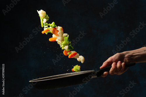Macro shot view of chef hand throws up mix of colored vegetables above the pan on dark blue background. Backstage of cooking meal. Frozen motion. Food banner concept. Cookbook illustration