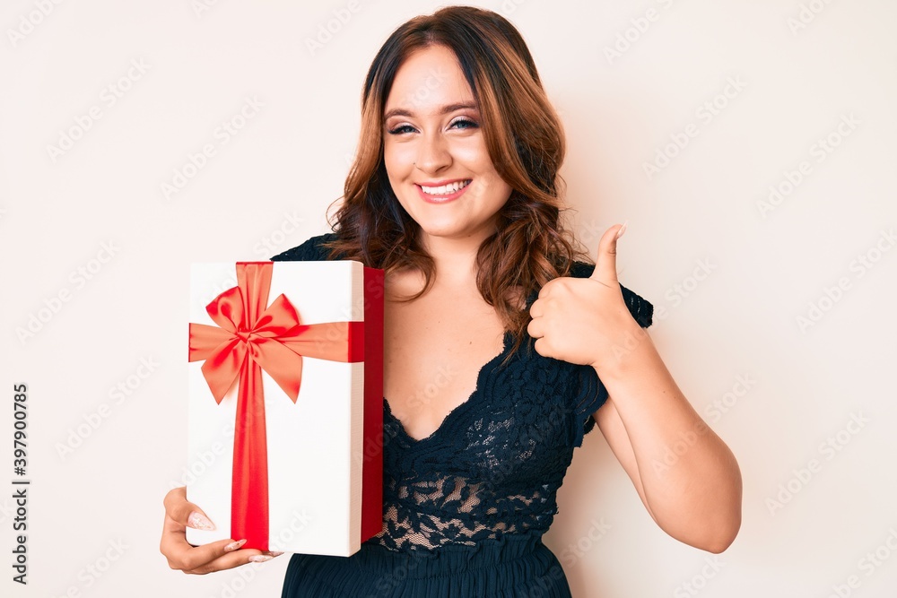 Young beautiful caucasian woman holding gift smiling happy and positive, thumb up doing excellent and approval sign Young beautiful caucasian woman holding gift smiling happy and positive, thumb up doing excellent and approval sign