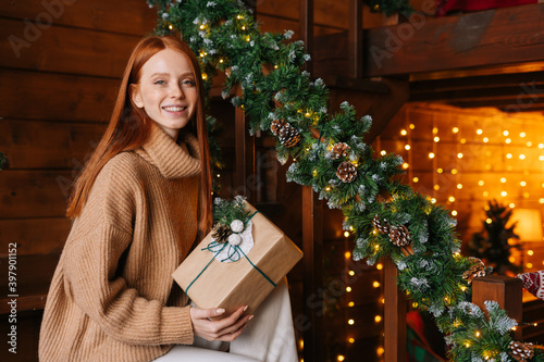 Happy redhead young woman holding gift box in hand received for Christmas at cozy living room with festive interior, looking at camera. Charming lady wearing winter sweater with holiday box at home.