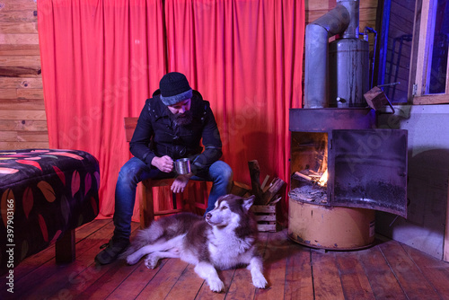 Bearded lumberjack is inside a log cabin drinking tea from a metal mug sitting next to his husky dog ​​enjoying the warmth provided by an old fireplace made from a rusty barrel with burning firewood