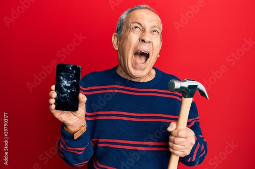 Senior hispanic man holding broken smartphone showing cracked screen and hammer angry and mad screaming frustrated and furious, shouting with anger looking up.
