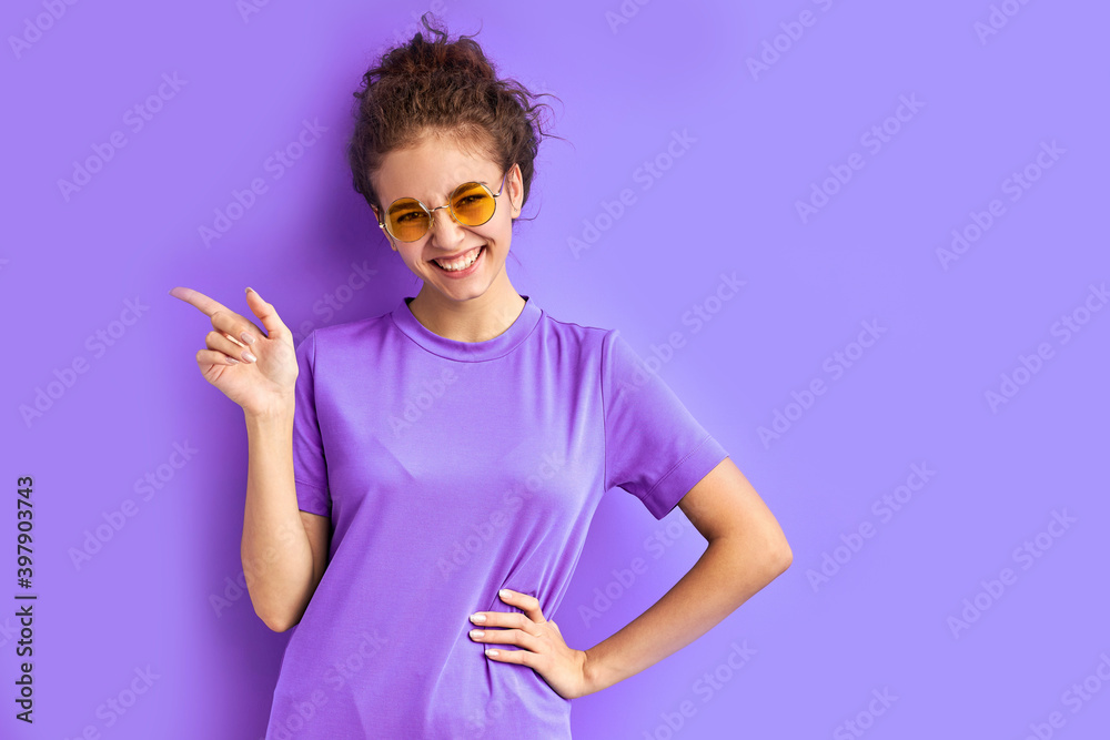 joyful teenager girl in t-shirt and sunglasses posing at camera, pointing fingers at side, index at left. isolated over purple background joyful teenager girl in t-shirt and sunglasses posing at camera, pointing fingers at side, index at left. isolated over purple background