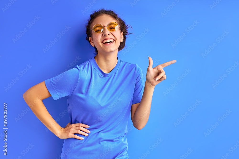 joyful teenager girl in t-shirt and sunglasses posing at camera, pointing fingers at side, index at right. isolated over blue background