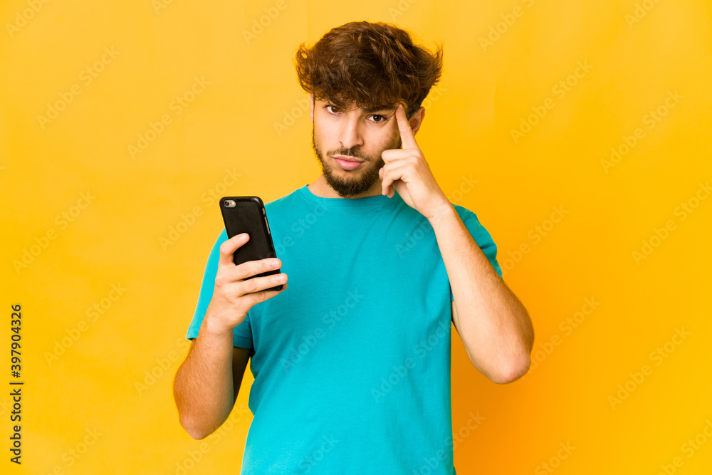 Young indian man holding a phone pointing temple with finger, thinking, focused on a task.
