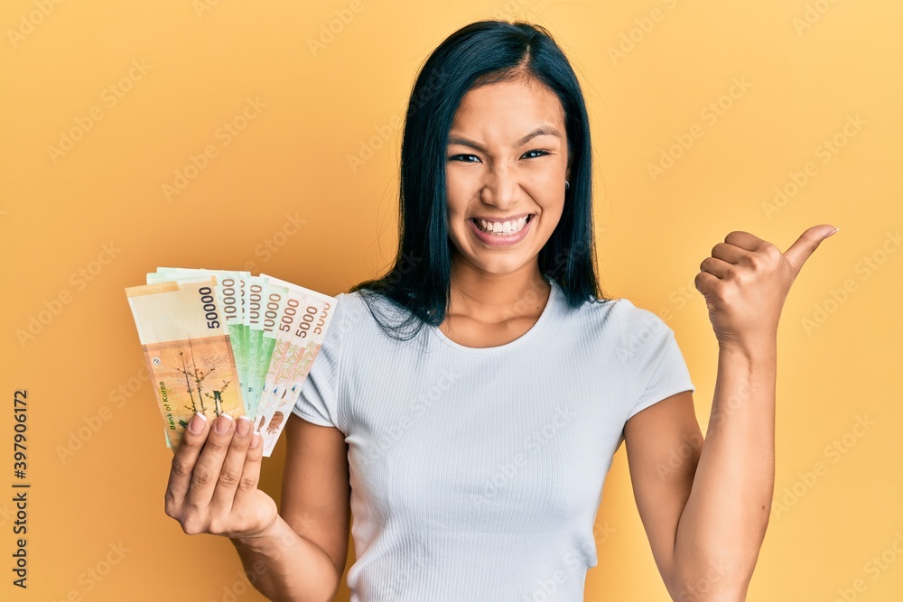 Beautiful hispanic woman holding south korean won banknotes pointing thumb up to the side smiling happy with open mouth Beautiful hispanic woman holding south korean won banknotes pointing thumb up to the side smiling happy with open mouth