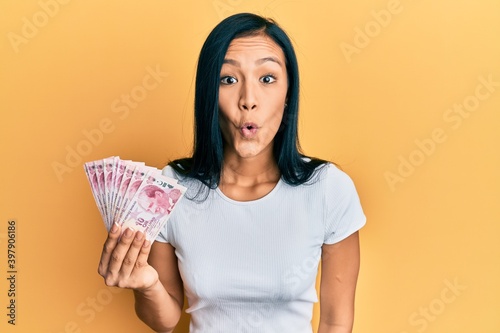 Beautiful hispanic woman holding 10 turkish lira banknotes scared and amazed with open mouth for surprise, disbelief face