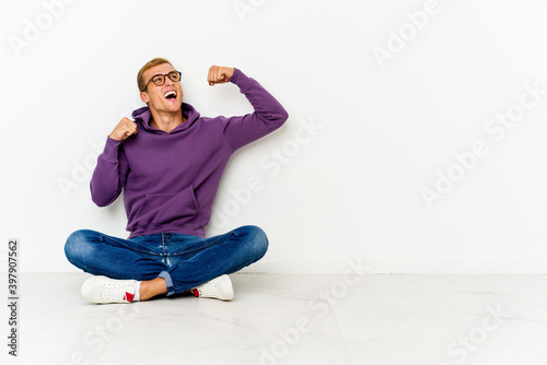 Young caucasian man sitting on the floor raising fist after a victory, winner concept.