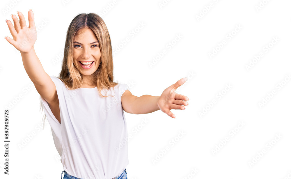 Beautiful caucasian woman with blonde hair wearing casual white tshirt looking at the camera smiling with open arms for hug. cheerful expression embracing happiness.