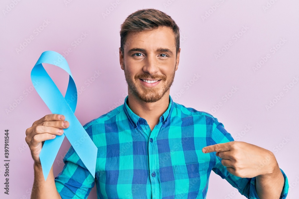 Handsome caucasian man holding blue ribbon pointing finger to one self smiling happy and proud Handsome caucasian man holding blue ribbon pointing finger to one self smiling happy and proud