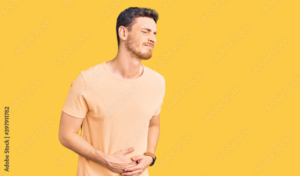 Handsome young man with bear wearing casual yellow tshirt with hand on stomach because nausea, painful disease feeling unwell. ache concept.
