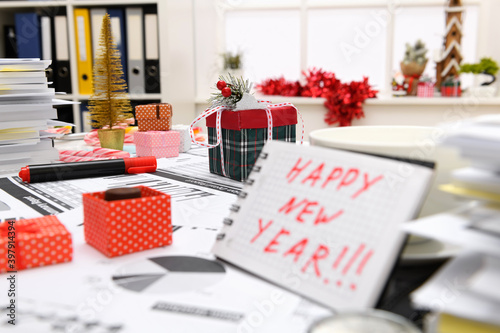 Business and holiday concept - office interior decorated with Christmas tree for celebrating, candies and other new year decoration. A notepad with happy new year text.