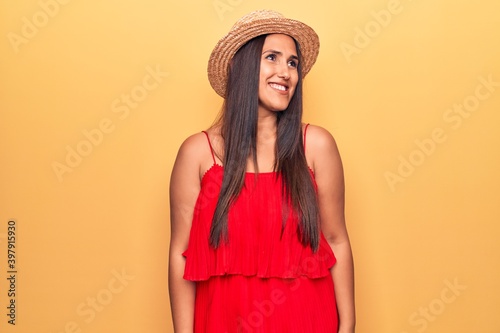 Young beautiful brunette woman wearing summer hat and dress looking away to side with smile on face, natural expression. laughing confident.