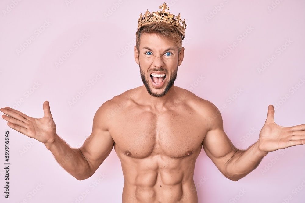 Young caucasian man shirtless wearing king crown celebrating victory with happy smile and winner expression with raised hands Young caucasian man shirtless wearing king crown celebrating victory with happy smile and winner expression with raised hands