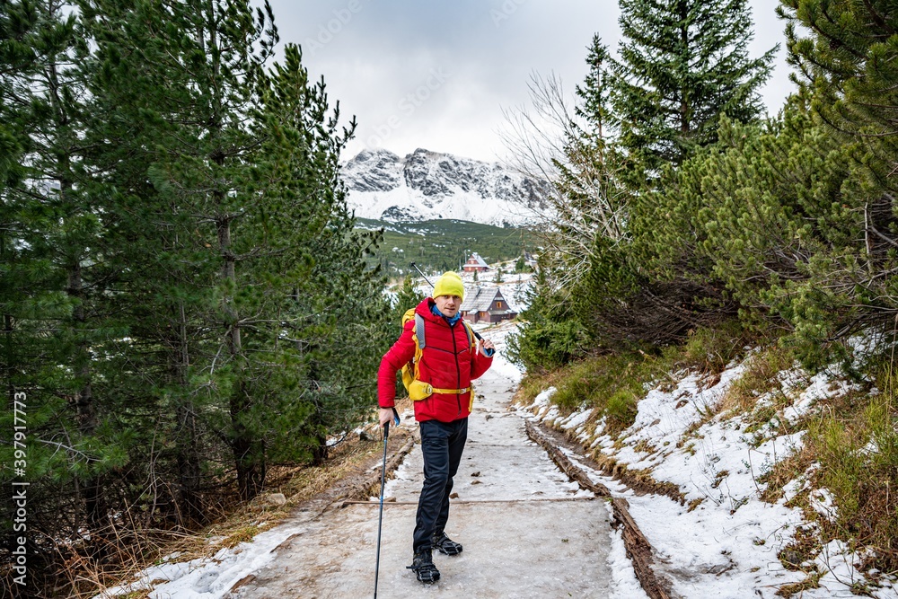 Hiker in a red down jacket, yellow hat and yellow backpack stands on a mountain trail with Nordic walking poles.