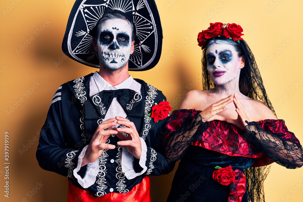 Young couple wearing mexican day of the dead costume over yellow hands together and fingers crossed smiling relaxed and cheerful. success and optimistic