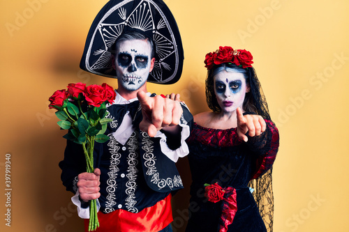 Young couple wearing mexican day of the dead costume holding roses pointing with finger to the camera and to you, confident gesture looking serious