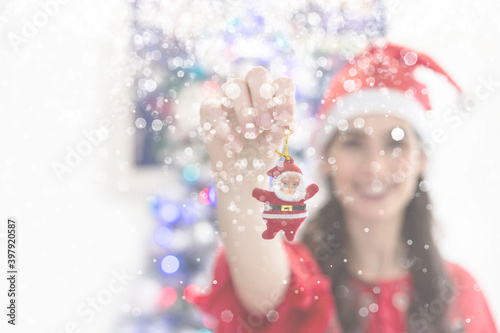 girl wearing christmas hat. smiling and holding santa claus teddy with tree and christmas lights background. christmas concept. snowing.