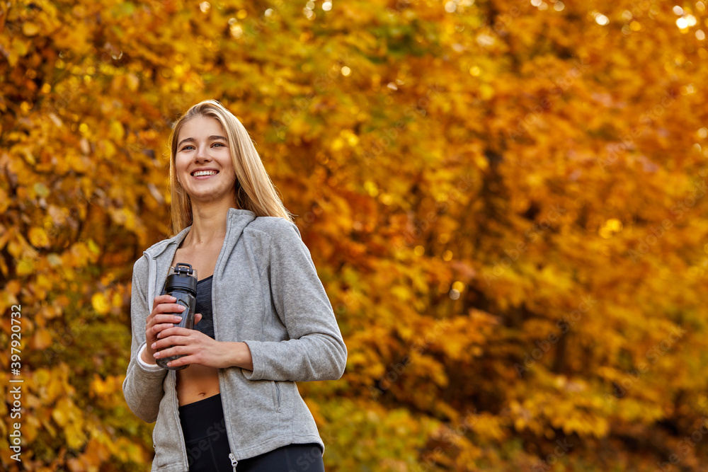 blonde female holding bottle of water in the forest, portrait. positive lady in sportswear have rest after running, jogging. at sunny autumn day blonde female holding bottle of water in the forest, portrait. positive lady in sportswear have rest after running, jogging. at sunny autumn day