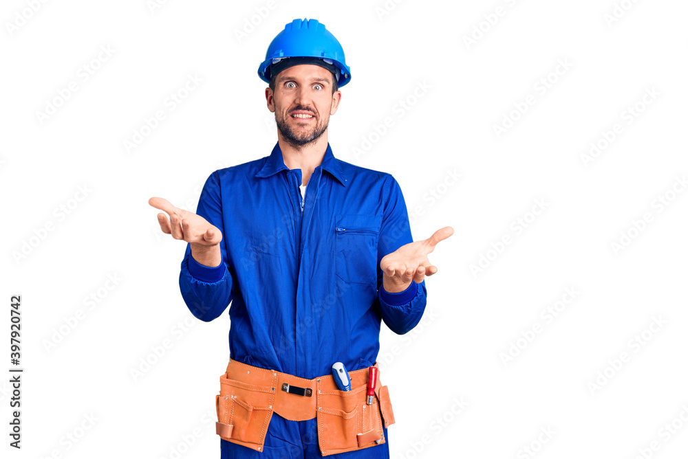 Young handsome man wearing worker uniform and hardhat smiling cheerful with open arms as friendly welcome, positive and confident greetings