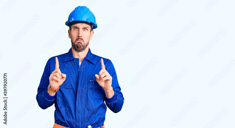 Young handsome man wearing worker uniform and hardhat pointing down looking sad and upset, indicating direction with fingers, unhappy and depressed. Young handsome man wearing worker uniform and hardhat pointing down looking sad and upset, indicating direction with fingers, unhappy and depressed.
