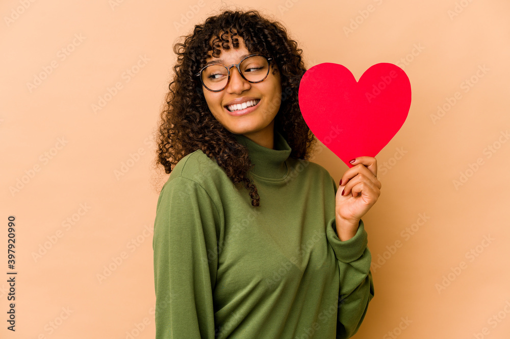 Young african american afro woman holding a valentines day heart looks aside smiling, cheerful and pleasant. Young african american afro woman holding a valentines day heart looks aside smiling, cheerful and pleasant.