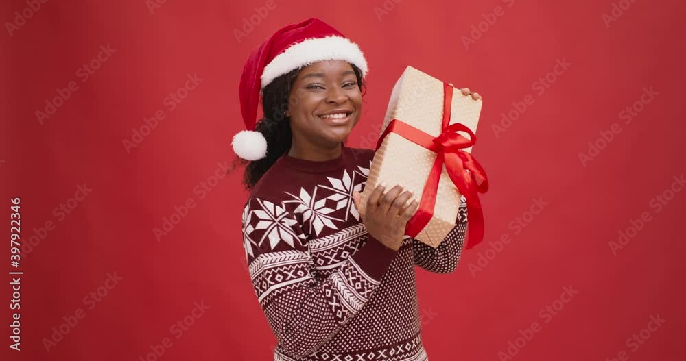 Curious black lady in santa hat shaking gift box, enjoying Christmas holiday, red background