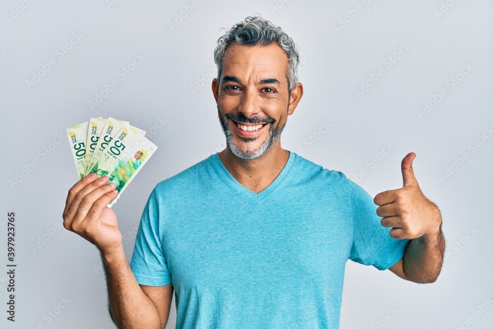 Middle age grey-haired man holding israel shekels banknotes smiling happy and positive, thumb up doing excellent and approval sign Middle age grey-haired man holding israel shekels banknotes smiling happy and positive, thumb up doing excellent and approval sign