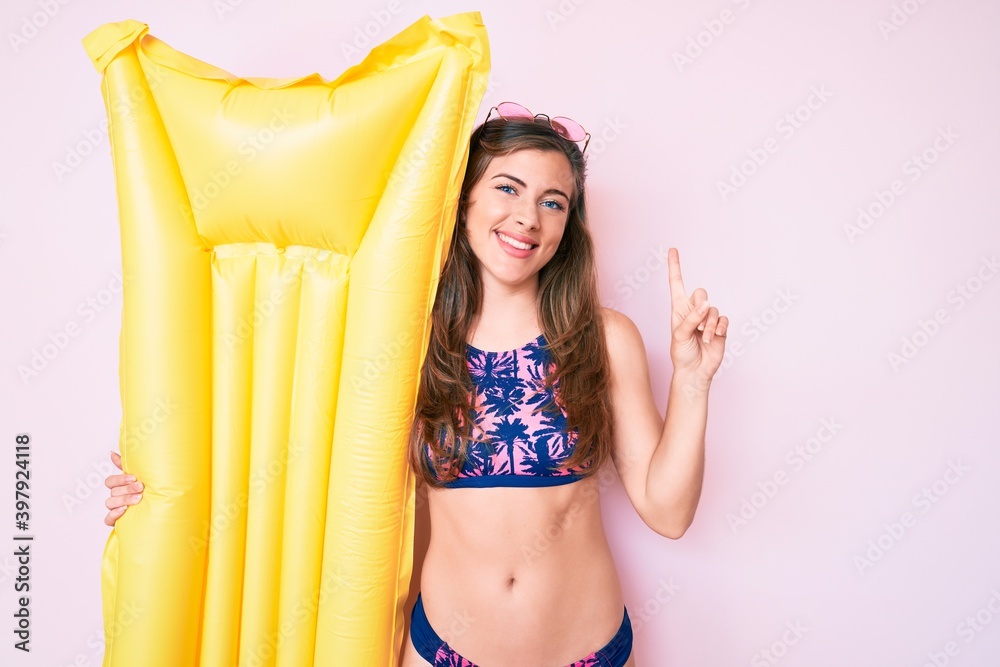 Obraz premium Beautiful young caucasian woman wearing bikini and holding summer matress float surprised with an idea or question pointing finger with happy face, number one