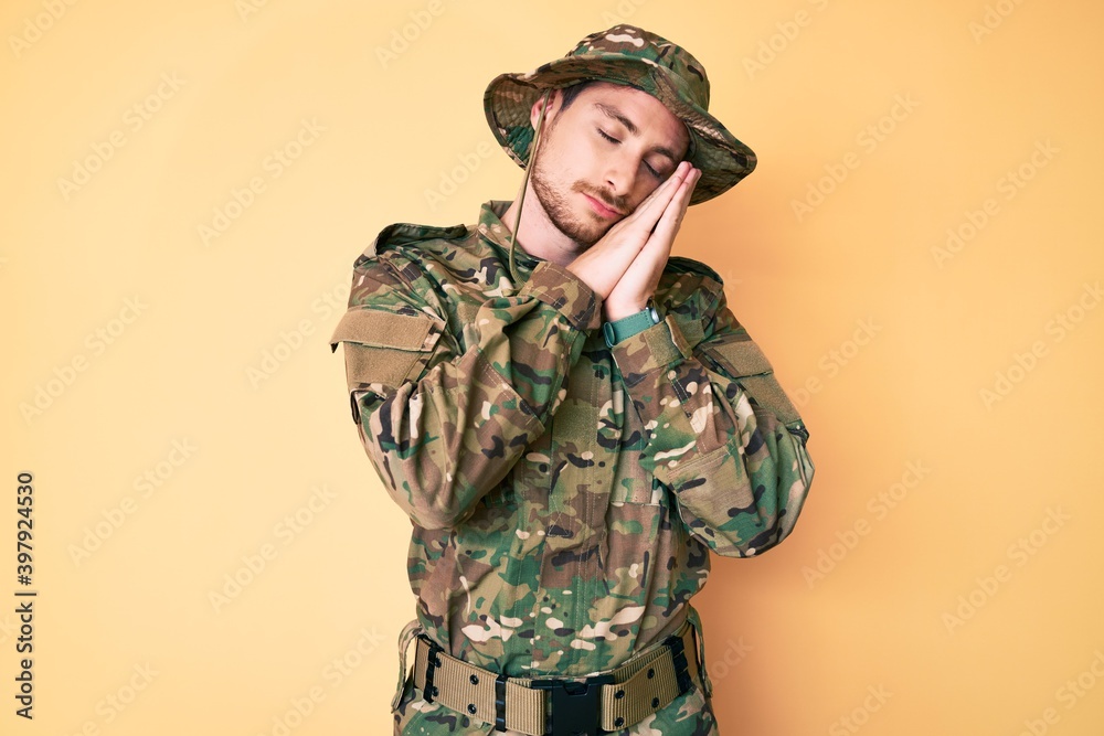 Young caucasian man wearing camouflage army uniform sleeping tired dreaming and posing with hands together while smiling with closed eyes. Young caucasian man wearing camouflage army uniform sleeping tired dreaming and posing with hands together while smiling with closed eyes.