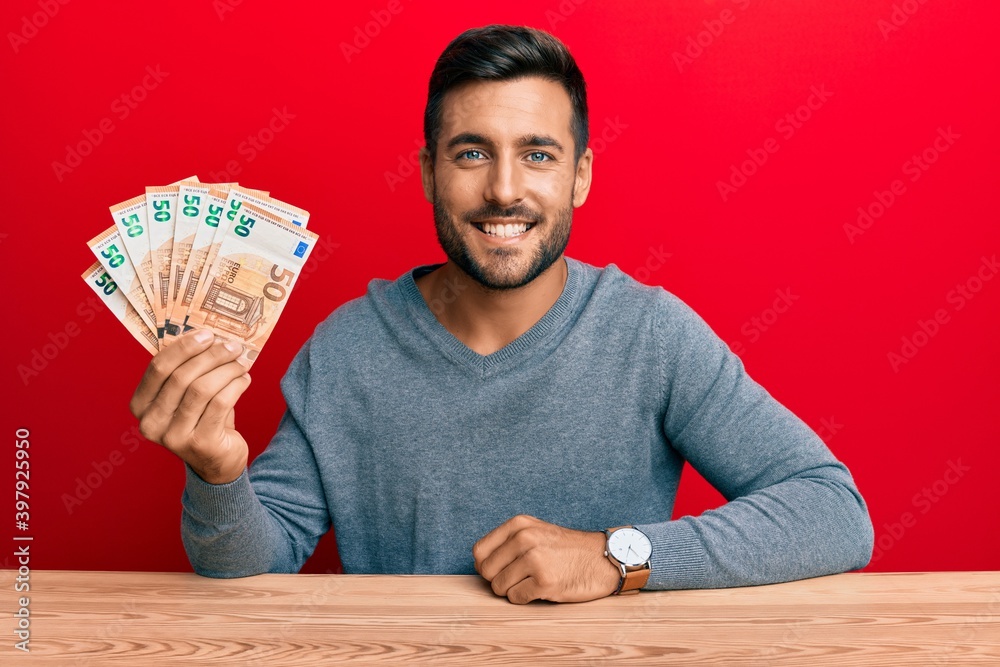 Handsome hispanic man holding bunch of 50 euro banknotes looking positive and happy standing and smiling with a confident smile showing teeth Handsome hispanic man holding bunch of 50 euro banknotes looking positive and happy standing and smiling with a confident smile showing teeth