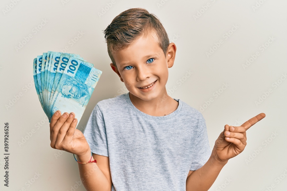 Adorable caucasian kid holding 100 brazilian real banknotes smiling happy pointing with hand and finger to the side Adorable caucasian kid holding 100 brazilian real banknotes smiling happy pointing with hand and finger to the side
