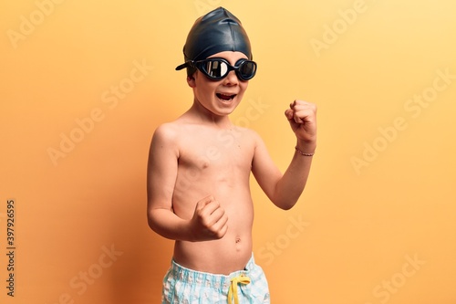 Cute blond kid wearing swimwear and swimmer glasses celebrating surprised and amazed for success with arms raised and eyes closed © Krakenimages.com