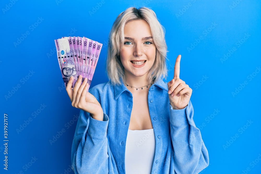 Young blonde girl holding philippine peso banknotes smiling with an idea or question pointing finger with happy face, number one