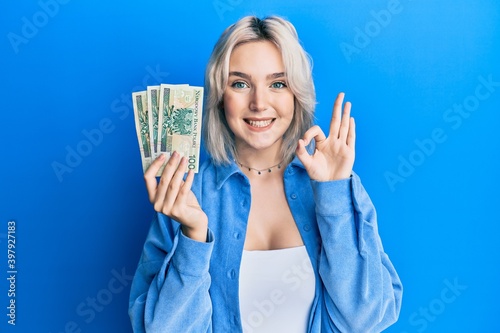 Young blonde girl holding 50 polish zloty banknotes doing ok sign with fingers, smiling friendly gesturing excellent symbol
