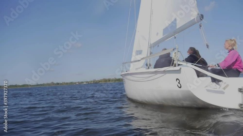 A young guy in sunglasses and two charming girls control the sail on a yacht during a trip on the river. Slow motion effect.