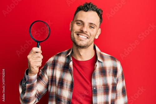 Young hispanic man holding magnifying glass looking for stain at clothes looking positive and happy standing and smiling with a confident smile showing teeth © Krakenimages.com
