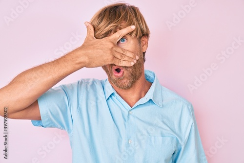 Young blond man wearing casual clothes peeking in shock covering face and eyes with hand, looking through fingers afraid