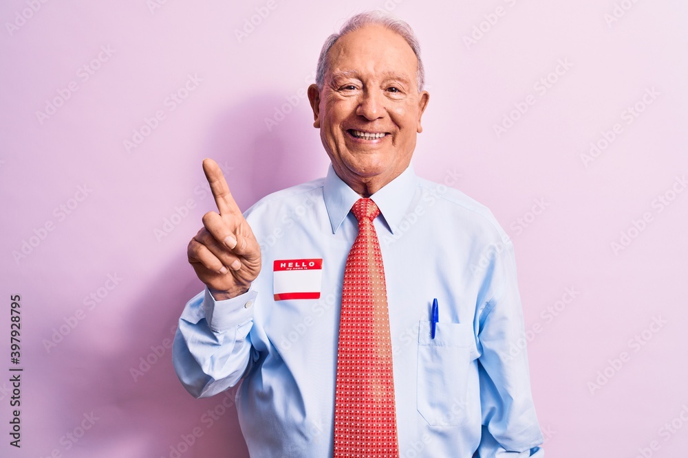 Senior handsome grey-haired businessman wearing tie and shirt with name presentation sticker smiling with an idea or question pointing finger up with happy face, number one Senior handsome grey-haired businessman wearing tie and shirt with name presentation sticker smiling with an idea or question pointing finger up with happy face, number one