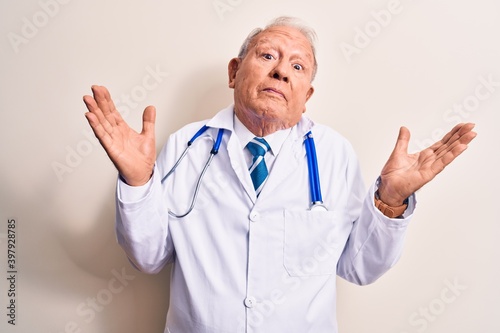 Senior grey-haired doctor man wearing coat and stethoscope standing over white background clueless and confused with open arms, no idea and doubtful face.