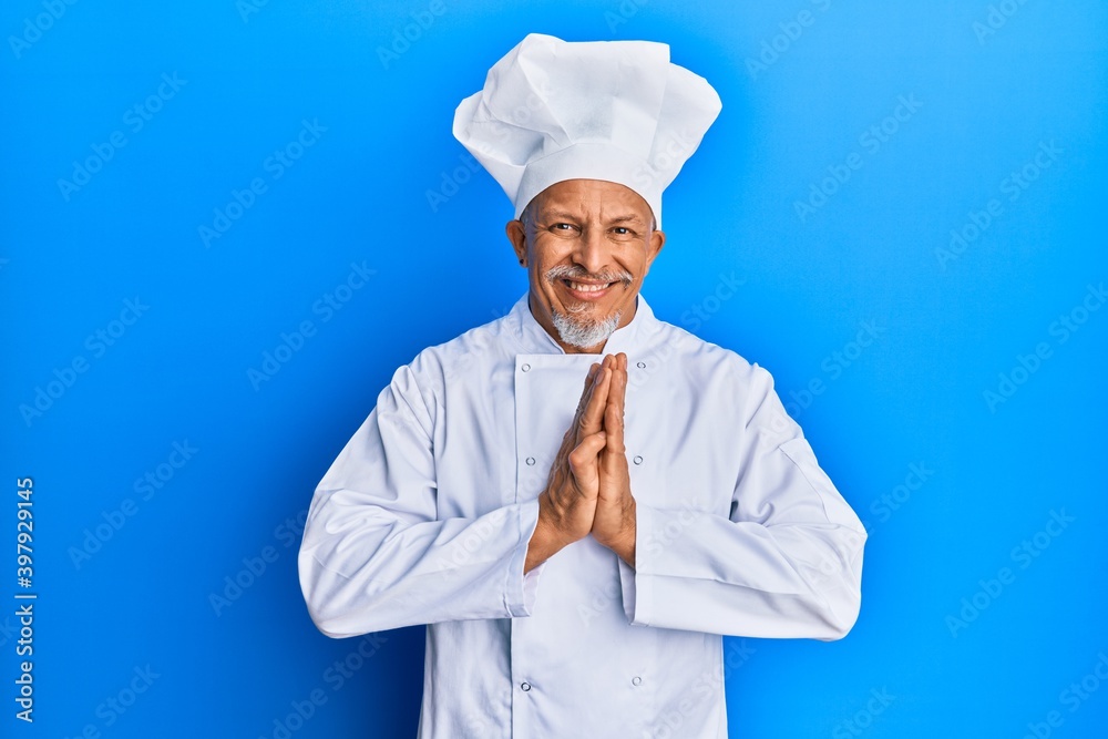Middle age grey-haired man wearing professional cook uniform and hat praying with hands together asking for forgiveness smiling confident.