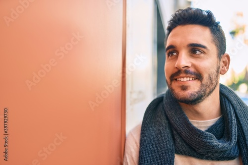 Young hispanic man smiling happy wearing scarf leaning on the wall at the city