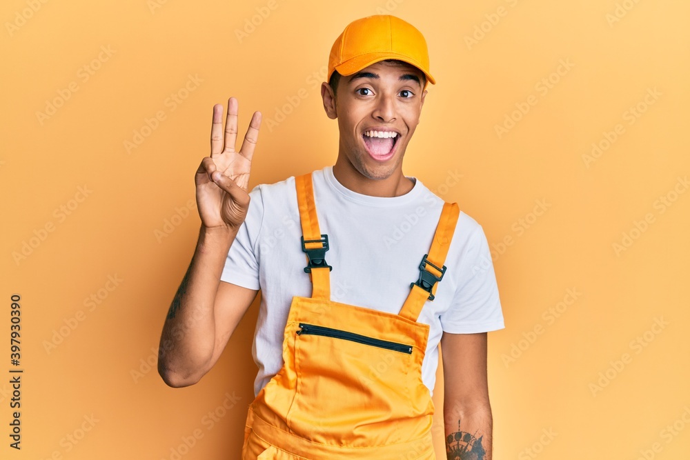 Young handsome african american man wearing handyman uniform over yellow background showing and pointing up with fingers number three while smiling confident and happy. Young handsome african american man wearing handyman uniform over yellow background showing and pointing up with fingers number three while smiling confident and happy.