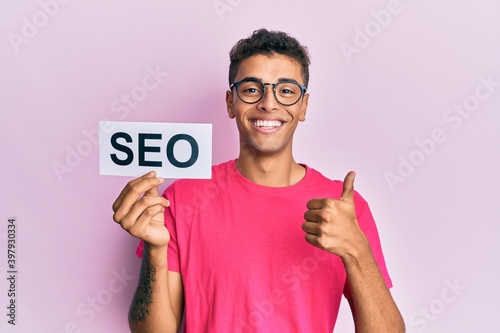 Young handsome african american man holding seo message paper smiling happy and positive, thumb up doing excellent and approval sign photo