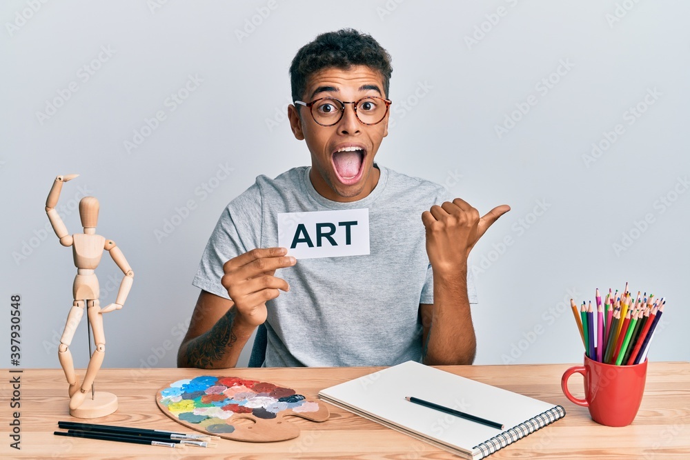 Young handsome african american man painter sitting at art studio holding art word pointing thumb up to the side smiling happy with open mouth Young handsome african american man painter sitting at art studio holding art word pointing thumb up to the side smiling happy with open mouth