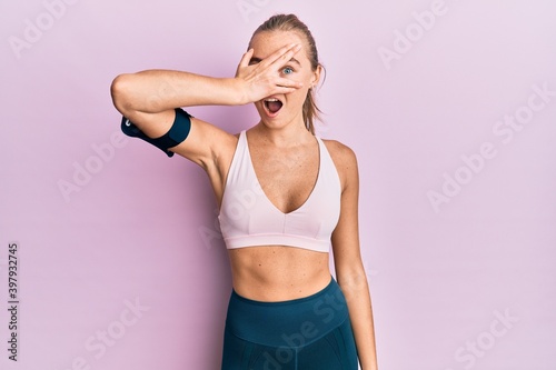 Beautiful blonde woman wearing sportswear and arm band peeking in shock covering face and eyes with hand, looking through fingers with embarrassed expression. © Krakenimages.com