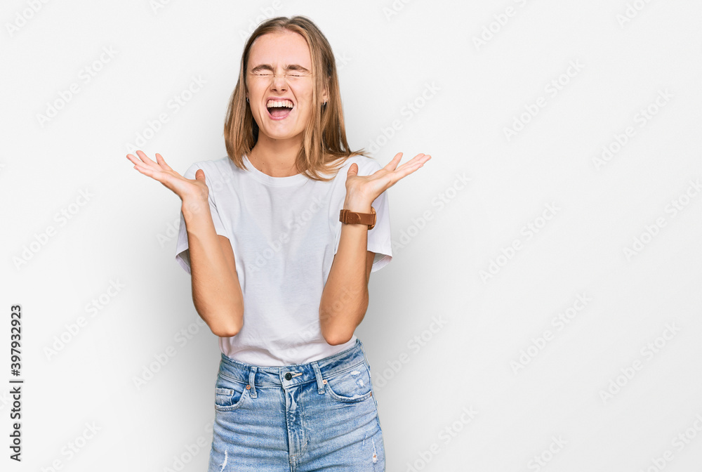 Beautiful young blonde woman wearing casual white t shirt celebrating mad and crazy for success with arms raised and closed eyes screaming excited. winner concept Beautiful young blonde woman wearing casual white t shirt celebrating mad and crazy for success with arms raised and closed eyes screaming excited. winner concept