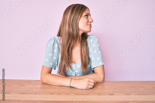 Young blonde girl wearing casual clothes sitting on the table looking to side, relax profile pose with natural face with confident smile.