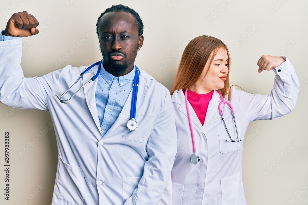 Young interracial couple wearing doctor uniform and stethoscope showing arms muscles smiling proud. fitness concept. Young interracial couple wearing doctor uniform and stethoscope showing arms muscles smiling proud. fitness concept.