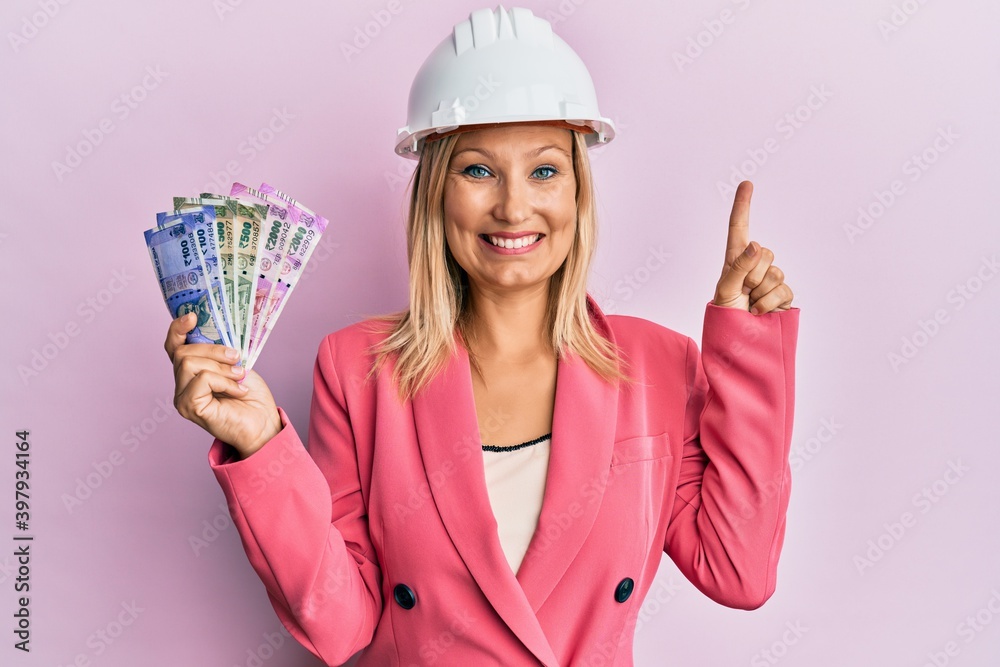 Beautiful middle age blonde woman wearing architect hardhat holding indian rupee smiling with an idea or question pointing finger with happy face, number one Beautiful middle age blonde woman wearing architect hardhat holding indian rupee smiling with an idea or question pointing finger with happy face, number one